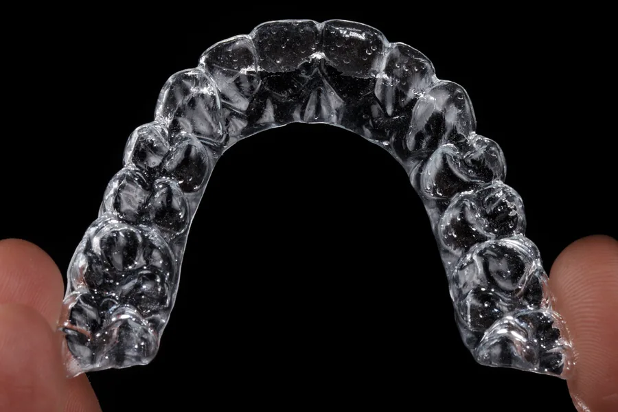 Alinhador transparente Invisalign visto de cima, segurado entre dois dedos sobre fundo preto